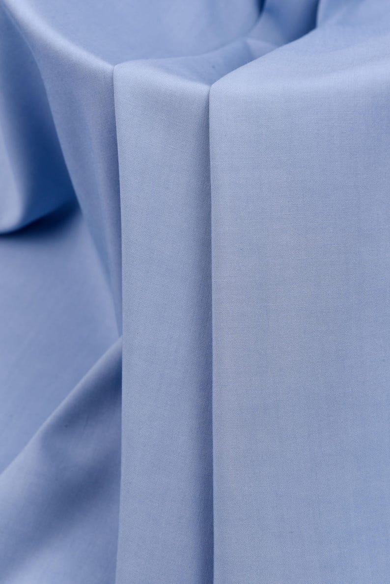May include: Close-up of a light blue fabric with a smooth texture. The fabric is folded in a way that creates a series of vertical lines.