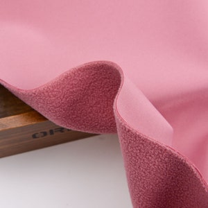 K&ouml;nnte beinhalten: Nahaufnahme eines rosa Fleece-Stoffs mit einer strukturierten, flauschigen Seite und einer glatten Seite. Der Stoff ist gefaltet und zeigt beide Texturen und Farben. Der Stoff wird wahrscheinlich f&uuml;r Kleidung oder Decken verwendet.