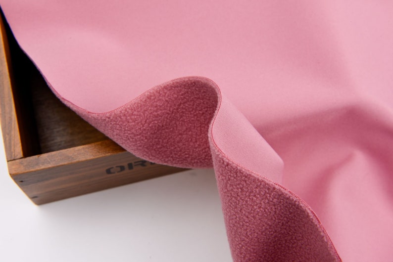 K&ouml;nnte beinhalten: Nahaufnahme eines rosa Fleece-Stoffs mit einer strukturierten, flauschigen Seite und einer glatten Seite. Der Stoff ist gefaltet und zeigt beide Texturen und Farben. Der Stoff wird wahrscheinlich f&uuml;r Kleidung oder Decken verwendet.