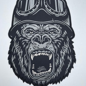 May include: Black and white embroidered patch of a gorilla wearing a leather aviator helmet and goggles. The gorilla is roaring with its mouth open, showing its teeth.