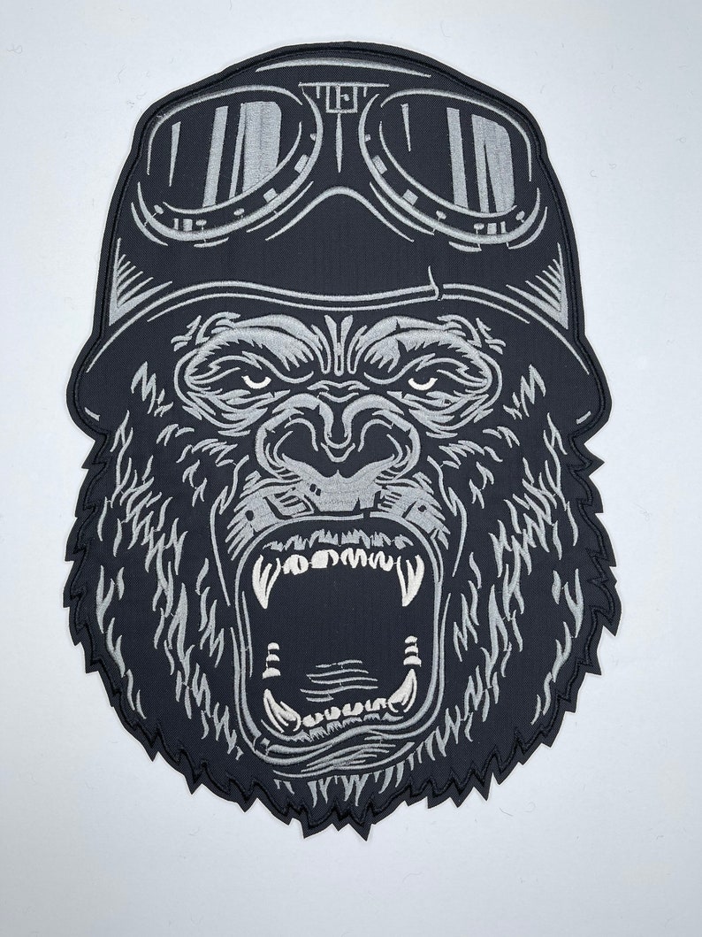 May include: Black and white embroidered patch of a gorilla wearing a leather aviator helmet and goggles. The gorilla is roaring with its mouth open, showing its teeth.