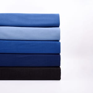 May include: A stack of folded fabric in shades of blue, ranging from light to dark. The colors include light blue, medium blue, navy, and black. The fabric appears to be a soft material, possibly cotton or linen.