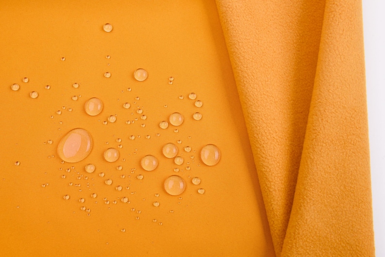 K&ouml;nnte beinhalten: Nahaufnahme eines goldgelben Stoffes mit Wassertropfen auf der Oberfl&auml;che. Ein Teil des Stoffes ist gefaltet und zeigt seine Textur. Die Tropfen sind unterschiedlich gro&szlig; und erzeugen einen dynamischen visuellen Effekt. Der Stoff wirkt weich.