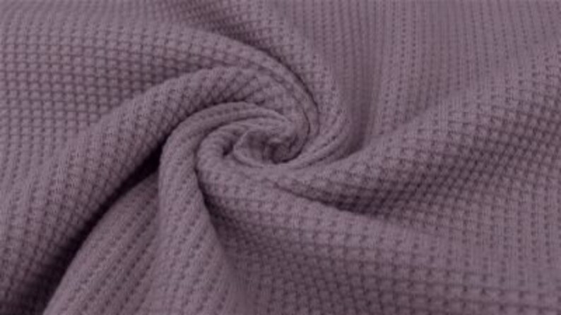 May include: Close-up of a swirl of mauve-colored waffle knit fabric. The fabric has a textured, grid-like pattern, with a soft, slightly blurred appearance. The folds and curves of the fabric create a sense of depth and movement.
