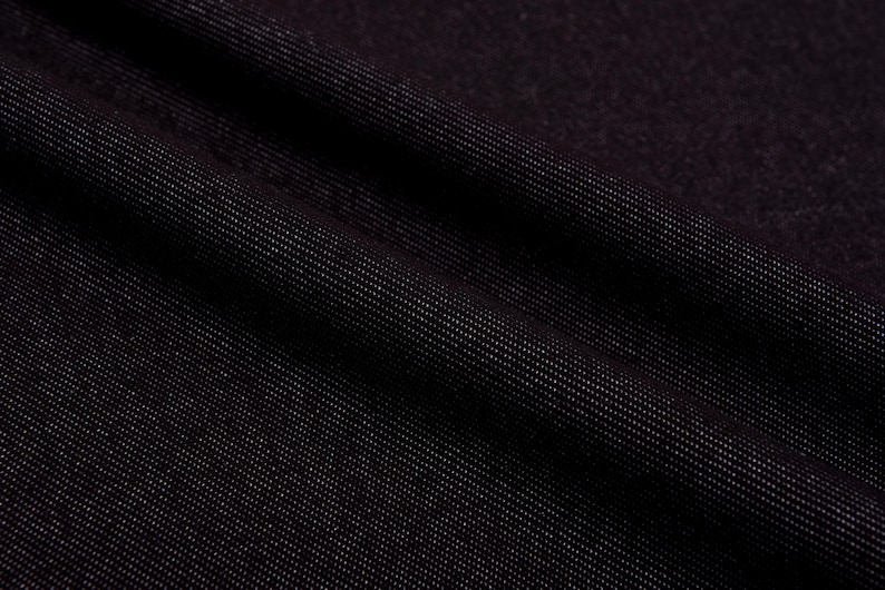 May include: Close-up of dark denim fabric with a subtle sheen. The fabric is a deep black color, with a diagonal weave pattern visible. The texture appears soft and pliable, with gentle folds and shadows.
