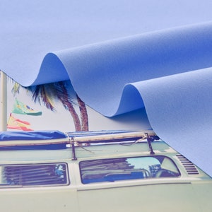 Może przedstawiać: Pr&oacute;bka jasnoniebieskiego materiału drapowana na vintage'owym vanie z deskami surfingowymi na dachu. Van jest zaparkowany przed palmami i plażą.