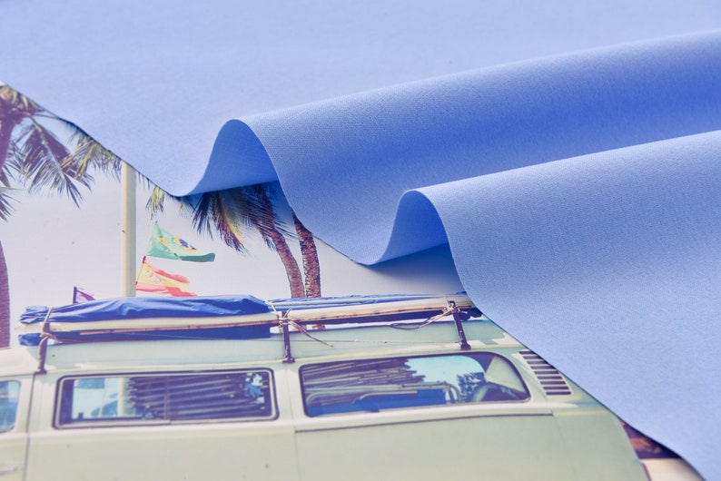 Może przedstawiać: Pr&oacute;bka jasnoniebieskiego materiału drapowana na vintage'owym vanie z deskami surfingowymi na dachu. Van jest zaparkowany przed palmami i plażą.