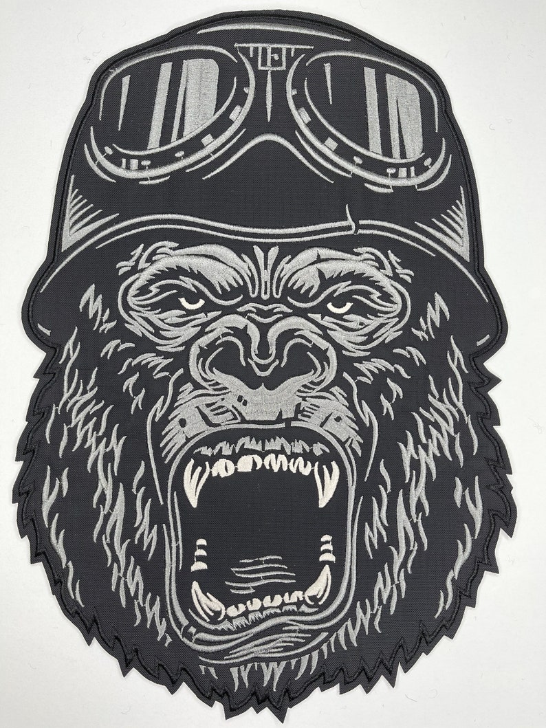 May include: A black and silver embroidered patch of a gorilla wearing a helmet and goggles. The gorilla is roaring with its mouth open, showing its teeth.