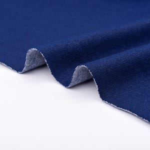 May include: Close-up of a piece of dark blue denim fabric. The fabric is folded, revealing a lighter, textured underside. The edges are raw and unfinished, showing the weave of the material. The denim has a classic, slightly worn appearance.
