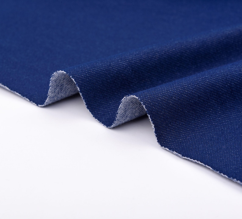 May include: Close-up of a piece of dark blue denim fabric. The fabric is folded, revealing a lighter, textured underside. The edges are raw and unfinished, showing the weave of the material. The denim has a classic, slightly worn appearance.