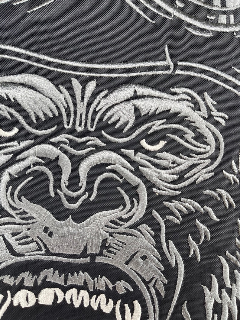 May include: Close-up of a silver embroidered gorilla face with an open mouth, showing teeth. The gorilla is on a black fabric background.