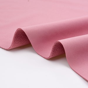 K&ouml;nnte beinhalten: Nahaufnahme eines gefalteten St&uuml;cks rosa Stoffes. Der Stoff hat eine weiche, samtige Textur auf der Oberseite und eine strukturierte, flauschige Unterseite. Die Farbe ist ein ged&auml;mpftes, staubiges Rosa, und der Stoff scheint ein mittleres Gewicht zu haben. Die Falten erzeugen Schatten und Tiefe.