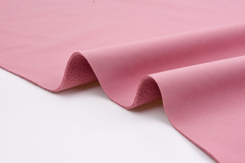 K&ouml;nnte beinhalten: Nahaufnahme eines gefalteten St&uuml;cks rosa Stoffes. Der Stoff hat eine weiche, samtige Textur auf der Oberseite und eine strukturierte, flauschige Unterseite. Die Farbe ist ein ged&auml;mpftes, staubiges Rosa, und der Stoff scheint ein mittleres Gewicht zu haben. Die Falten erzeugen Schatten und Tiefe.