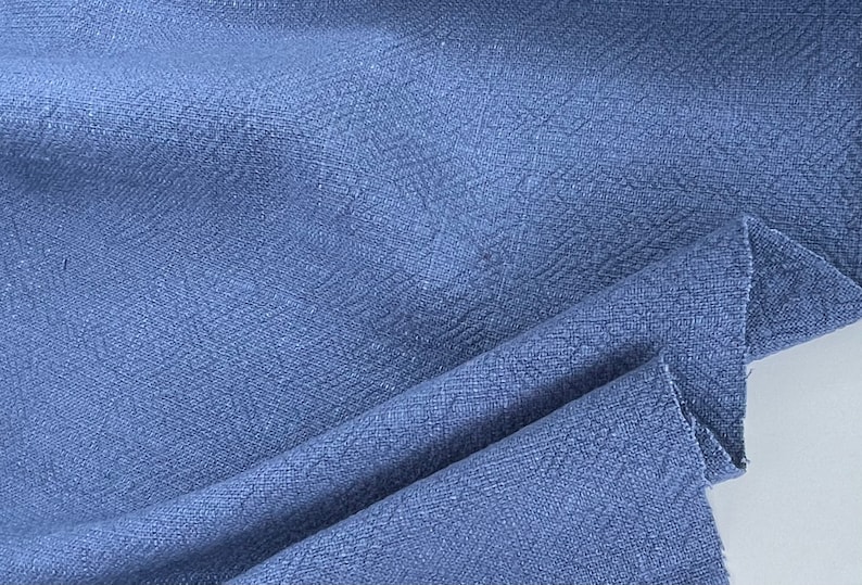 May include: Close-up of a blue textured fabric with a subtle herringbone pattern. The fabric is folded and draped, showcasing its soft and drapey texture.