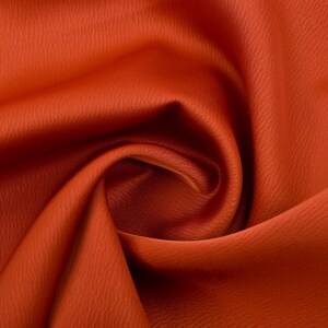 Puede incluir: Primer plano de una tela naranja vibrante con una superficie texturizada. El material est&aacute; drapeado y doblado, creando sombras suaves y reflejos. El color de la tela es consistente en toda la imagen.