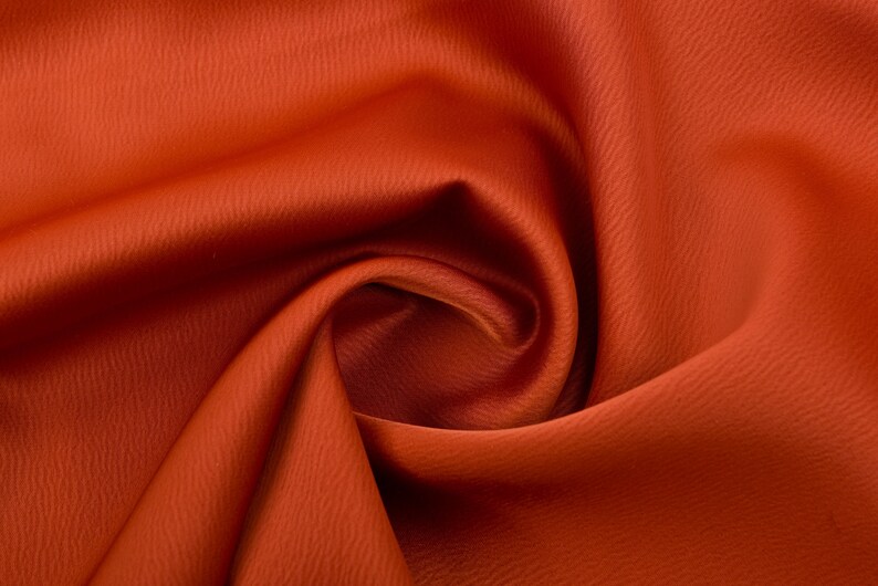 Puede incluir: Primer plano de una tela naranja vibrante con una superficie texturizada. El material est&aacute; drapeado y doblado, creando sombras suaves y reflejos. El color de la tela es consistente en toda la imagen.