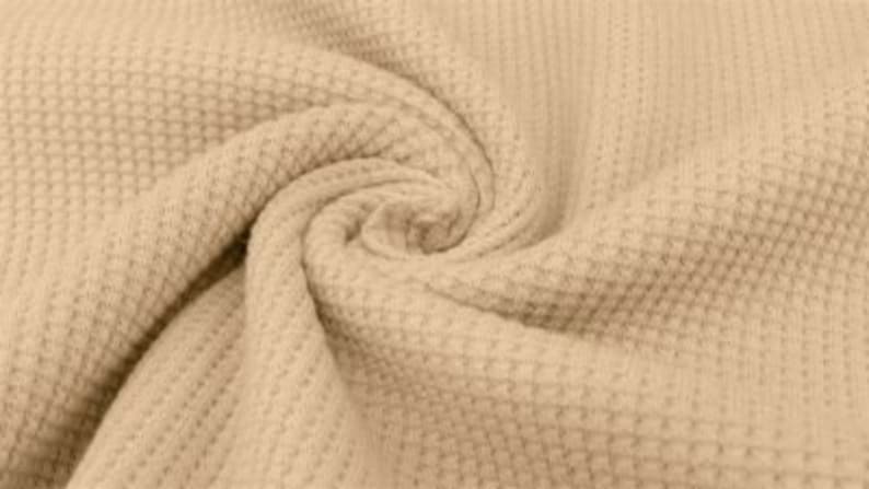 May include: Close-up of a beige waffle-knit fabric, tightly swirled to show texture. The fabric has a repeating grid pattern, creating a textured surface. The neutral color and detailed weave suggest a cozy textile.