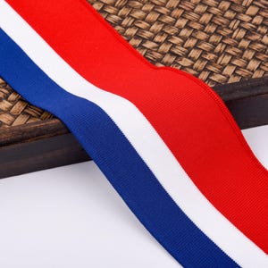 May include: A close-up of a wide, ribbed ribbon featuring three vertical stripes. The stripes are in the colors of red, white, and blue. The ribbon is lying on a woven, brown surface.