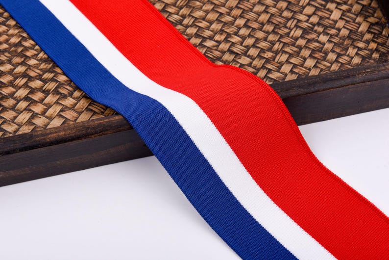 May include: A close-up of a wide, ribbed ribbon featuring three vertical stripes. The stripes are in the colors of red, white, and blue. The ribbon is lying on a woven, brown surface.