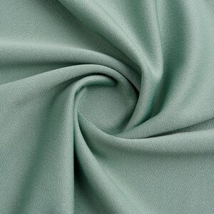 Pode incluir: Um close-up de um tecido macio de cor verde-s&aacute;lvia com uma textura lisa. O tecido est&aacute; dobrado e drapeado, criando um padr&atilde;o de redemoinho.