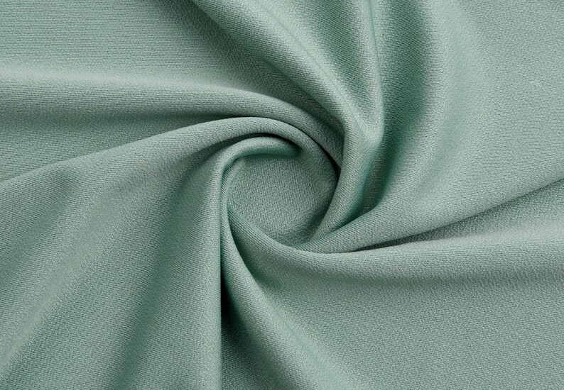 Pode incluir: Um close-up de um tecido macio de cor verde-s&aacute;lvia com uma textura lisa. O tecido est&aacute; dobrado e drapeado, criando um padr&atilde;o de redemoinho.