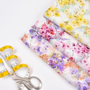 Peut inclure: Gros plan d'un m&egrave;tre ruban jaune et de ciseaux argent&eacute;s sur une surface blanche. Trois morceaux de tissu &agrave; motifs floraux sont empil&eacute;s les uns sur les autres. Le tissu a un aspect aquarelle avec des fleurs roses, violettes et jaunes.