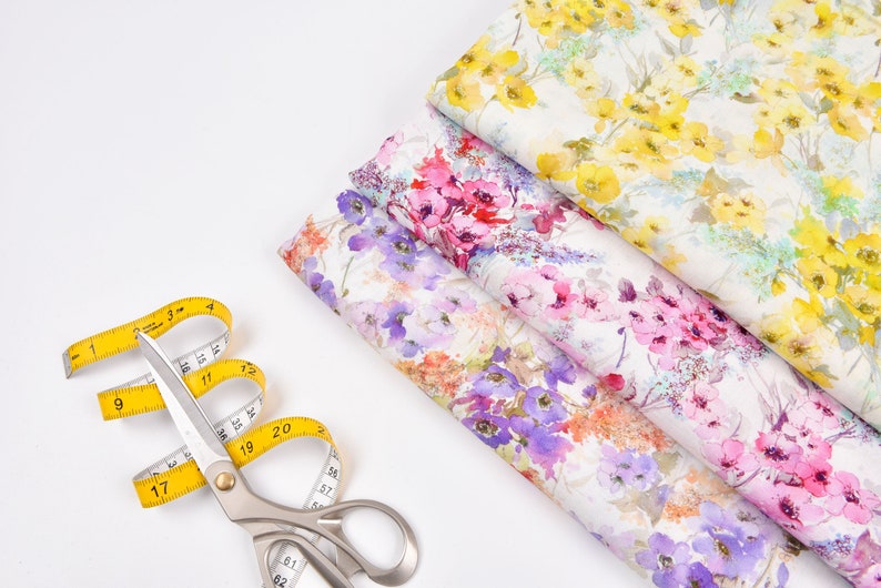 Peut inclure: Gros plan d'un m&egrave;tre ruban jaune et de ciseaux argent&eacute;s sur une surface blanche. Trois morceaux de tissu &agrave; motifs floraux sont empil&eacute;s les uns sur les autres. Le tissu a un aspect aquarelle avec des fleurs roses, violettes et jaunes.
