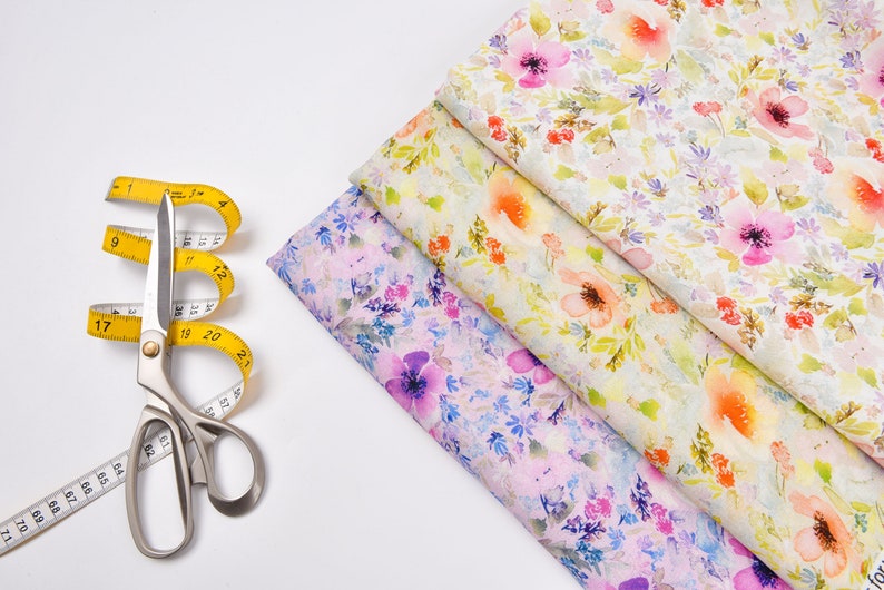 Peut inclure: Un ruban &agrave; mesurer jaune et des ciseaux argent&eacute;s sur une surface blanche, &agrave; c&ocirc;t&eacute; de trois morceaux de tissu avec des motifs floraux dans des tons de rose, jaune et blanc.