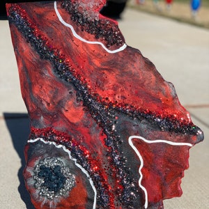 May include: A red, black and white resin art piece shaped like the state of Georgia. The piece features a glittery, abstract design with white lines and a black and silver centre.