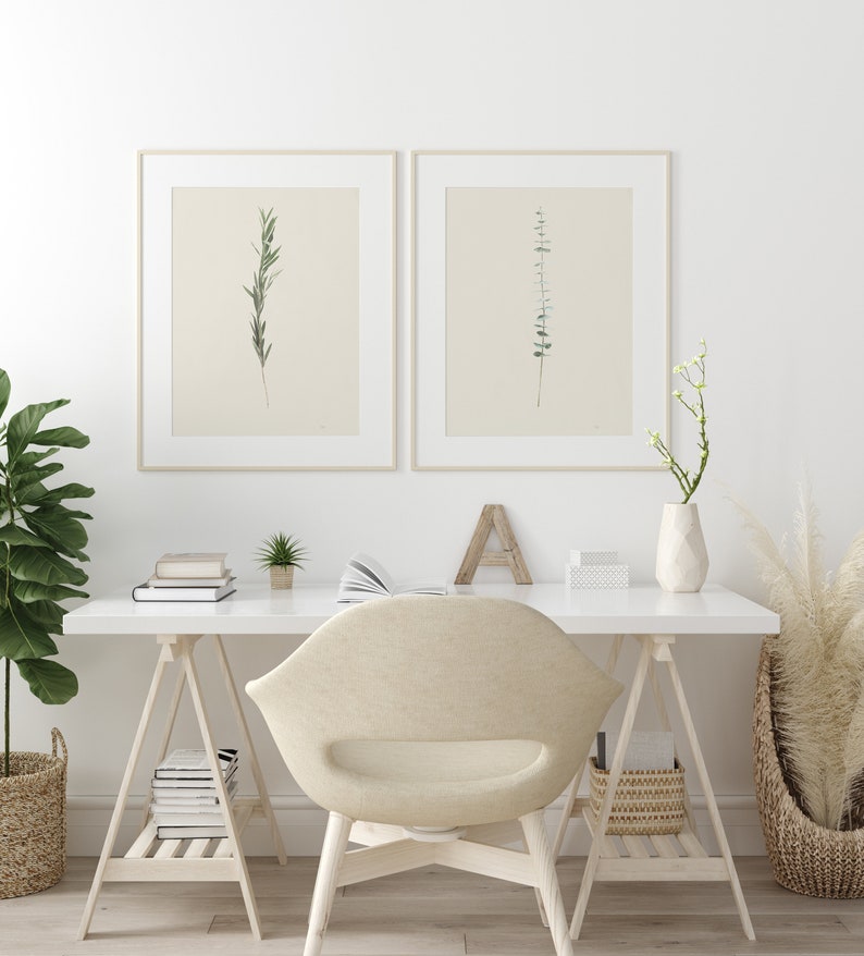 May include: A white desk with a white chair and two framed botanical prints of green leaves on a white background. The desk is on wooden sawhorse legs and has a white vase with white flowers on the right side. There are books and a small potted plant on the desk.