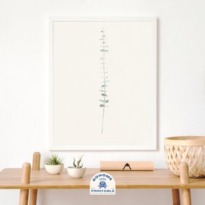 May include: A framed print of a single sprig of eucalyptus leaves in shades of green against a white background. The print is displayed on a wooden table with a woven basket, a straw hat, and two small potted plants.