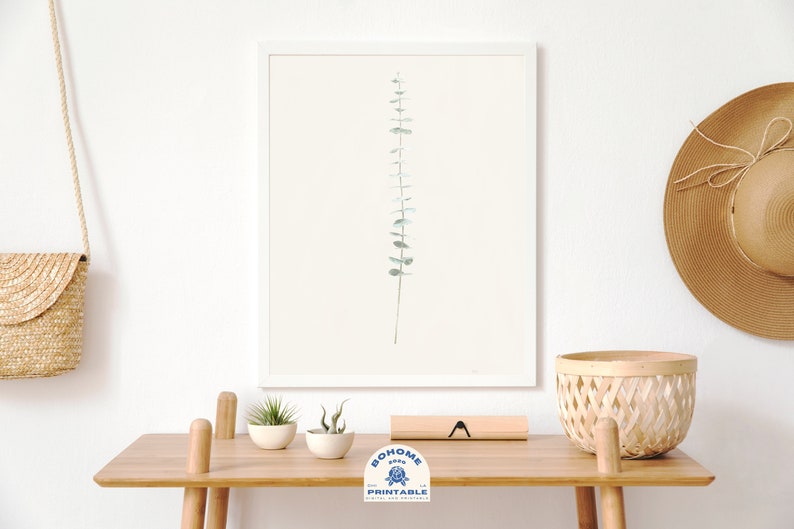 May include: A framed print of a single sprig of eucalyptus leaves in shades of green against a white background. The print is displayed on a wooden table with a woven basket, a straw hat, and two small potted plants.