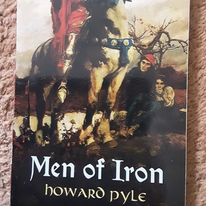 May include: A book cover with an illustration of a knight on a white horse. The knight is wearing red and gold armor and is holding a spear. The horse is wearing a blue and gold saddle. The book title is "Men of Iron" by Howard Pyle.