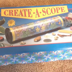 May include: A blue box with the words "CREATE-A-SCOPE" in yellow and red. The box contains a kaleidoscope with a floral design and small objects. The kaleidoscope is cylindrical and has a clear tube. The box is decorated with a floral pattern.