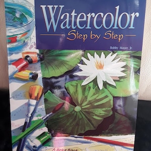 May include: A book titled "Watercolor Step by Step" with a blue cover. The cover features a watercolor painting of a white water lily, lily pads, and paintbrushes. The book is published by A Beka Book.
