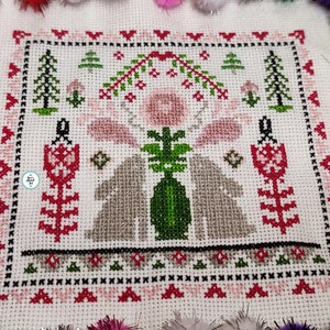 Op de afbeelding: Kruissteekborduurwerk met een centraal ontwerp van twee grijze konijnen die een groene vaas flankeren. Het ontwerp bevat roze bloemen, groene bomen en rode en zwarte randdetails op een witte achtergrond. Het stuk is omgeven door kleurrijke pompons.