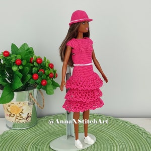 May include: A doll wearing a handmade, bright pink crochet dress with a tiered skirt and matching hat. The dress has a white belt and the doll is wearing white sneakers. The background includes a green placemat and a small plant.