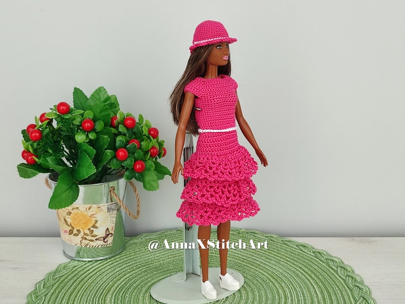 May include: A doll wearing a handmade, bright pink crochet dress with a tiered skirt and matching hat. The dress has a white belt and the doll is wearing white sneakers. The background includes a green placemat and a small plant.