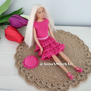 May include: A doll wearing a handmade, hot pink crochet outfit, including a top, tiered skirt, and matching hat. The doll is seated on a round, woven placemat. The outfit has a white belt. Pink sandals complete the ensemble.