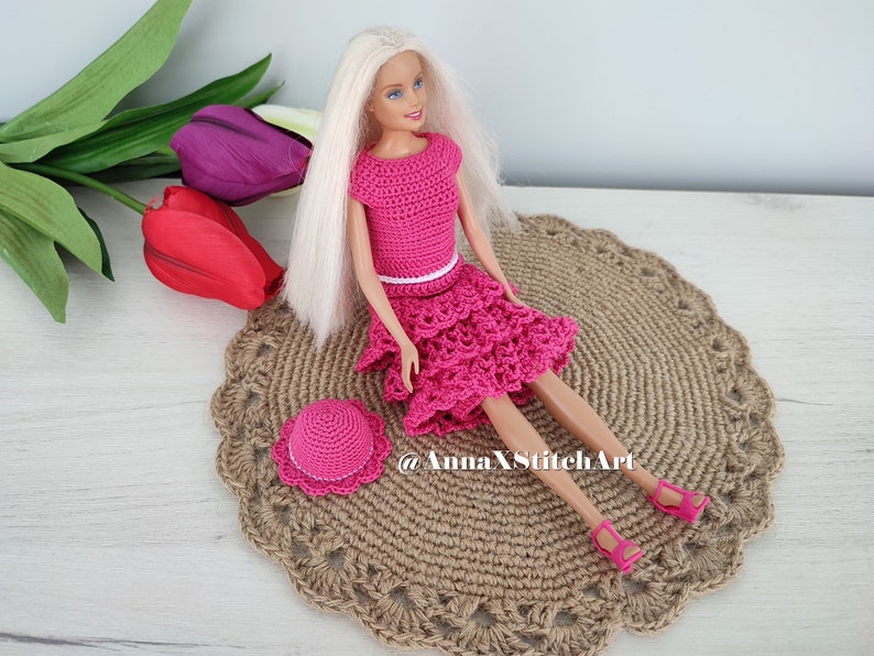 May include: A doll wearing a handmade, hot pink crochet outfit, including a top, tiered skirt, and matching hat. The doll is seated on a round, woven placemat. The outfit has a white belt. Pink sandals complete the ensemble.