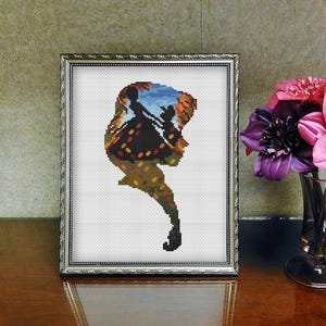 May include: Framed cross-stitch art featuring a silhouette of a figure with butterfly wings. The artwork uses a variety of colors, including orange, brown, blue, and green. The frame is silver and the artwork is displayed on a wooden surface.