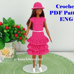 May include: A doll wearing a handmade, bright pink crochet outfit, including a top, tiered skirt, and hat. The outfit is accented with a white belt and trim. Text in the image reads "Crochet PDF Pattern ENG".