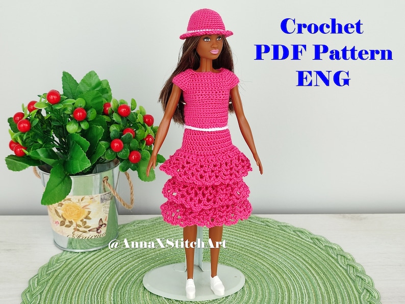 May include: A doll wearing a handmade, bright pink crochet outfit, including a top, tiered skirt, and hat. The outfit is accented with a white belt and trim. Text in the image reads "Crochet PDF Pattern ENG".