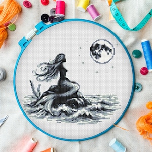 May include: A circular blue embroidery hoop containing white fabric with a cross-stitch design of a mermaid sitting on a rock in the ocean under a full moon. The mermaid and moon are rendered in shades of grey and black thread on the white fabric.