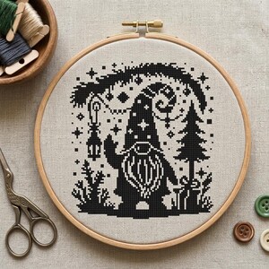 May include: A black and white cross-stitch design of a gnome holding a lantern, set in a wooden embroidery hoop. The gnome is surrounded by stars, a tree, and other decorative elements. A wooden bowl of thread, scissors, and buttons are also visible.