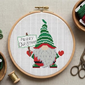 May include: A cross-stitch embroidery in a wooden hoop features a gnome holding a sign that reads "Merry Christmas." The gnome has a green and white striped hat, a white beard, and red accents. The embroidery is surrounded by sewing supplies.