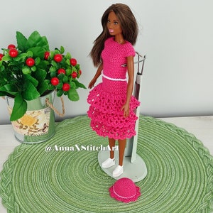 May include: A doll wearing a handmade, bright pink crochet outfit, including a top and tiered skirt, with a matching hat and white sneakers. The outfit has white trim. The doll is standing on a metal stand, with a green placemat underneath. A small potted plant is in the background.