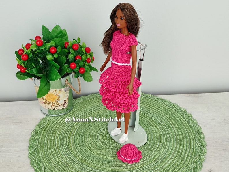 May include: A doll wearing a handmade, bright pink crochet outfit, including a top and tiered skirt, with a matching hat and white sneakers. The outfit has white trim. The doll is standing on a metal stand, with a green placemat underneath. A small potted plant is in the background.