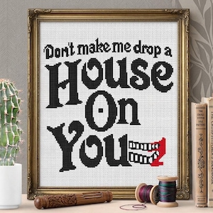 May include: Cross-stitch art in a gold frame with the text "Don't make me drop a House On You." The black text is on a white background. A red mouth with teeth is at the bottom. A cactus, books, and spools of thread are in the background.