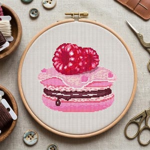 Op de afbeelding: Een kruissteekborduurring met een roze macaron met een framboos erop. Omringd door houten spoelen met draad, knopen en gouden scharen, wat duidt op een knutselthema.